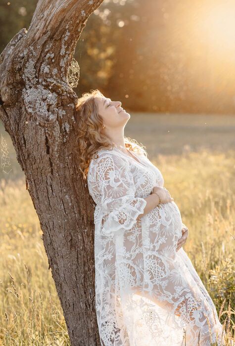 Fotoshooting mit Babybauch in der Natur im Nürnberger Land Schwangere in Spitzenkleid lehnt am Baum